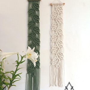 Puede incluir: Dos tapices de macramé de pared con un patrón de hojas. El de la izquierda es verde y el de la derecha es blanco. Ambos tapices tienen una varilla de madera en la parte superior y una franja en la parte inferior. El texto "Kieuhandmade Original items from real people" está en la parte inferior de la imagen.