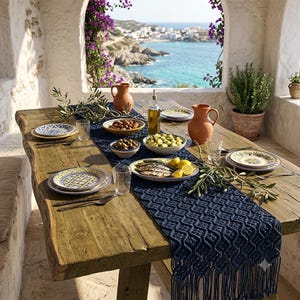 May include: A rustic wooden dining table features a dark blue macrame table runner with fringe. The table is set with patterned ceramic plates, clear glasses, two terracotta pitchers, a glass oil bottle, and bowls containing olives and fish.