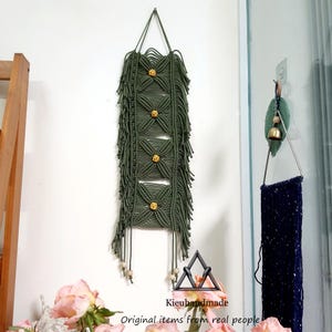 May include: Olive green macrame wall hanging with a geometric design. The handmade piece features a series of diamond-shaped patterns, accented with small, round, golden-colored beads. The bottom has fringe and wooden beads. The text "Kieuhandmade" is at the bottom.