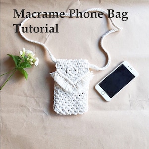 May include: A white macrame phone bag with a fringe detail and a long strap. The bag is laying on a brown surface with a white smartphone next to it.