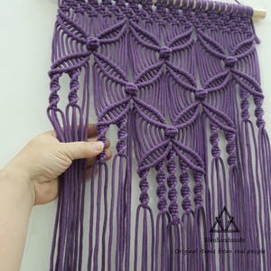 May include: A handcrafted macrame wall hanging in a vibrant purple hue. The design features intricate knotting patterns, including floral-like motifs. The piece is suspended from a natural wooden dowel. The text "Kieuhandmade Original items from real people" is visible.