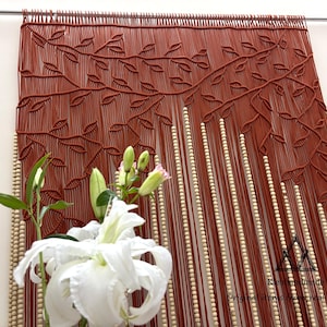 May include: A rust-coloured macrame wall hanging featuring a detailed leaf pattern woven into the top section, with vertical strands accented by light-coloured wooden beads. A small black triangular logo with the text Kleihandmade is visible.