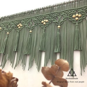 May include: A green macrame wall hanging with wooden beads and tassels. The hanging is made with a knotted design and has a fringe at the bottom.