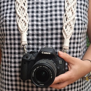 Puede incluir: Una correa de cámara de macramé blanca con una cámara Canon EOS 700D negra con un objetivo de 18-55 mm.
