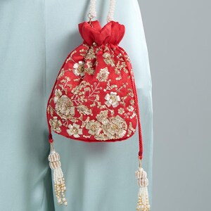 Può includere: Una borsa rossa con coulisse e ricami floreali dorati. La borsa presenta un cordino rosso e nappe di perline bianche. La borsa è sospesa da un cordino bianco. Un piccolo accessorio decorativo.