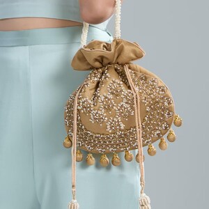 Könnte beinhalten: Ein goldfarbener Beutel mit aufwendiger weißer und goldener Perlenarbeit. Die Tasche hat einen Perlenhenkel und dekorative Quasten. Der Beutel wird vor einem hellblauen Hintergrund gehalten.