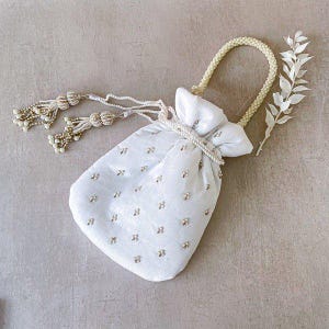 Puede incluir: Un bolso blanco con cordón, asa de cuentas y borlas decorativas. La bolsa está adornada con pequeños adornos dispersos. Una ramita de hojas blancas descansa junto a la bolsa, creando una estética delicada y elegante.