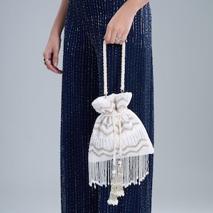 Könnte beinhalten: Weißer Beutel mit goldenen und silbernen Stickereien. Die Tasche hat einen Perlenhenkel und eine lange Perlenquaste. Die Tasche wird vor einem dunkelblauen Pailletten-Jumpsuit gehalten.