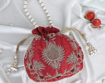 Op de afbeelding: Een rode tas met trekkoord, gouden borduursel en een met parels bezette handgreep. De tas heeft een bloemenontwerp en is afgewerkt met parels. Twee rode decoratieve ornamenten staan op de achtergrond.