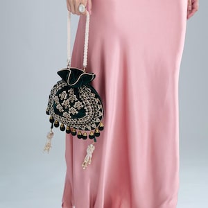 Puede incluir: Un bolso con cordón de terciopelo verde oscuro con intrincados bordados dorados y borlas de cuentas. El bolso tiene un asa con cuentas de perlas y un ribete dorado. El bolso se sostiene contra un vestido rosa.
