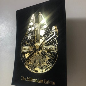 May include: A black poster with a gold foil print of the Millennium Falcon spaceship. The text "The Millennium Falcon" is printed in gold foil at the bottom of the poster.