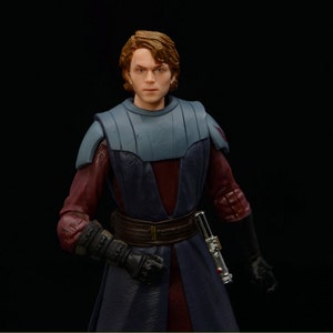 May include: A Star Wars action figure of Anakin Skywalker in his Jedi Knight attire. He is wearing a dark blue and burgundy robe with a brown belt and a lightsaber on his hip. He has brown hair and a serious expression on his face.