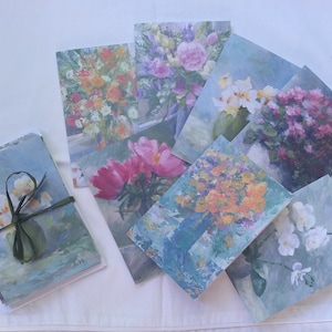 May include: A set of floral art greeting cards. The cards display colourful paintings of diverse flowers in vases, with a soft, painterly style. A stack of cards is tied with a green ribbon.