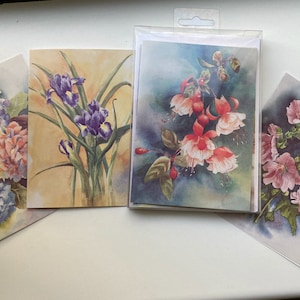 May include: Set of four greeting cards featuring watercolour paintings of various flowers. The cards feature a purple iris, a pink fuchsia, a pink hydrangea, and a pink hollyhock.