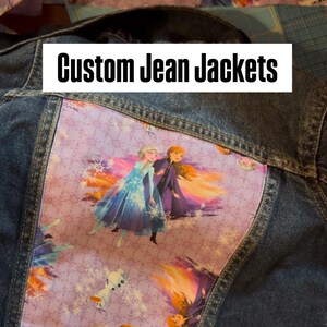 May include: Custom denim jacket with a purple panel featuring characters from the film Frozen. The text "Custom Jean Jackets" is displayed in a white rectangle. A unique piece of clothing.