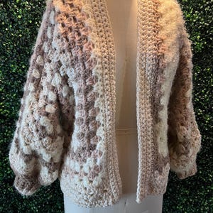 May include: A crocheted cardigan in shades of beige and brown with a textured pattern. The cardigan has a long, open front and long sleeves.