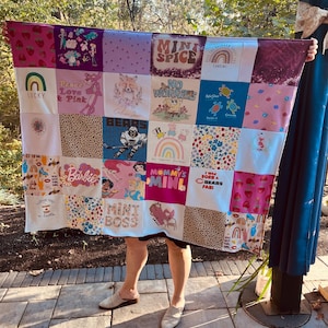 May include: A patchwork quilt made of various fabric squares. The squares feature colorful designs, including rainbows, cartoon characters, and text like "Lucky," "Barbie," and "Mommy's Mini." The quilt is held up by a person.