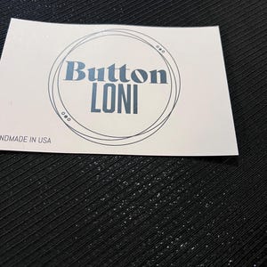 Könnte beinhalten: Eine weiße Karte mit einem schwarzen Kreis und dem Text "Button Loni" im Kreis. Der Text "Handmade in USA" ist unter dem Kreis gedruckt.