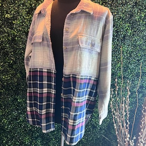 May include: A two-toned flannel shirt displayed on a mannequin. The top half is a light gray and lavender plaid, while the bottom half features a contrasting plaid in navy, black, and pink. The shirt has two chest pockets and a button-down front.