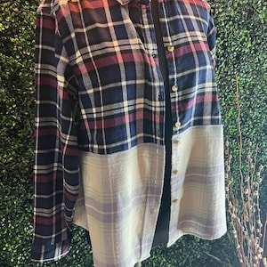 May include: A button-down flannel shirt in a patchwork design. The top half features a navy, burgundy, and white plaid pattern, while the bottom half is a light beige and lavender plaid. The shirt is displayed on a mannequin.