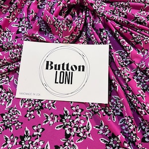 May include: Pink fabric with a white and black floral print. The fabric is folded and a white card with the text "Button Loni Handmade in USA" is on top of the fabric.