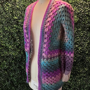 May include: A crocheted cardigan with a long open front and long sleeves. The cardigan is made with a multi-colored yarn in shades of purple, pink, and blue.