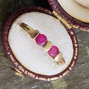 May include: A gold ring featuring two round, deep pink rubies set in a row, displayed inside a small, open, dark burgundy-colored circular jewelry box with a cream-colored interior lining and gold-toned trim along the edges of the lid.