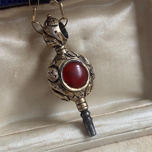 Könnte beinhalten: Antiker goldfarbener Anhänger mit einem roten Edelstein in der Mitte und aufwendigen floralen Details. Der Anhänger hängt an einer zarten Goldkette. Präsentiert in einer Vintage-Schmuckschatulle in Blau und Creme.
