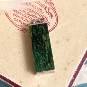 Può includere: Un ciondolo rettangolare con un'ansa argentata e una vivace pietra verde marmorizzata. La pietra ha una base verde scuro con venature verde chiaro. Il ciondolo è posto su uno sfondo di carta vintage con testo.