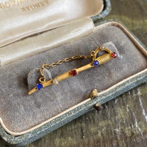 Könnte beinhalten: Eine goldfarbene Schwertbrosche mit einer dekorativen Kette, besetzt mit kleinen runden blauen, roten und klaren Steinen. Der Schwertgriff ist gerippt und die Klinge ist mit abwechselnd farbigen Edelsteinen entlang der Länge verziert.