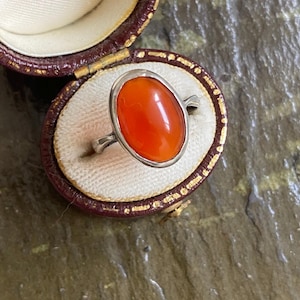 May include: A silver ring featuring a large, smooth, oval-shaped orange stone set in a simple metal bezel. The ring is displayed inside a small, oval-shaped, dark red vintage-style jewelry box with a cream-colored fabric interior lining.
