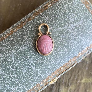 Puede incluir: Un colgante de oro con un escarabajo tallado en una piedra rosa de forma ovalada. El colgante tiene una pequeña anilla dorada redondeada en la parte superior para engancharlo a una cadena. La piedra muestra líneas grabadas detalladas de un escarabajo.