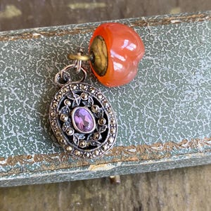 May include: A pendant featuring an oval silver-toned frame decorated with small dark grey stones and a central oval purple stone. Attached to the top is a small, rounded, translucent orange bead with a brass-coloured metal cap and a jump ring.