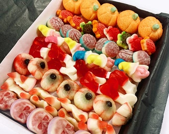 Puede incluir: Una caja blanca llena de caramelos gomosos de colores con temática de Halloween. Los caramelos tienen forma de calabazas, cerebros, ojos, calaveras, huesos y gusanos.