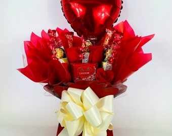 May include: A red heart-shaped balloon sits atop a red gift basket filled with assorted chocolates and a white bow. The basket is decorated with red tissue paper and a red ribbon.