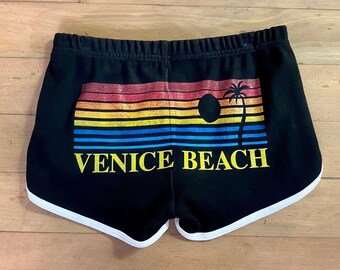 Może przedstawiać: Czarne spodenki sportowe z białym wykończeniem. Spodenki mają kolorowy wzór zachodu słońca z palmą i napisem "VENICE BEACH" w kolorze żółtym.