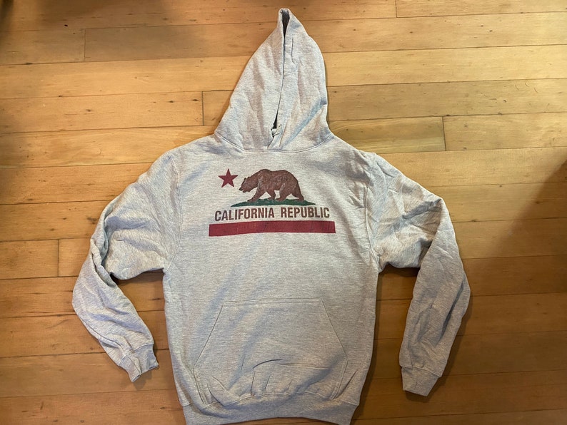 May include: A gray hooded sweatshirt with a graphic of the California Republic flag featuring a brown bear and a red star. The text "CALIFORNIA REPUBLIC" is printed in red below the bear.