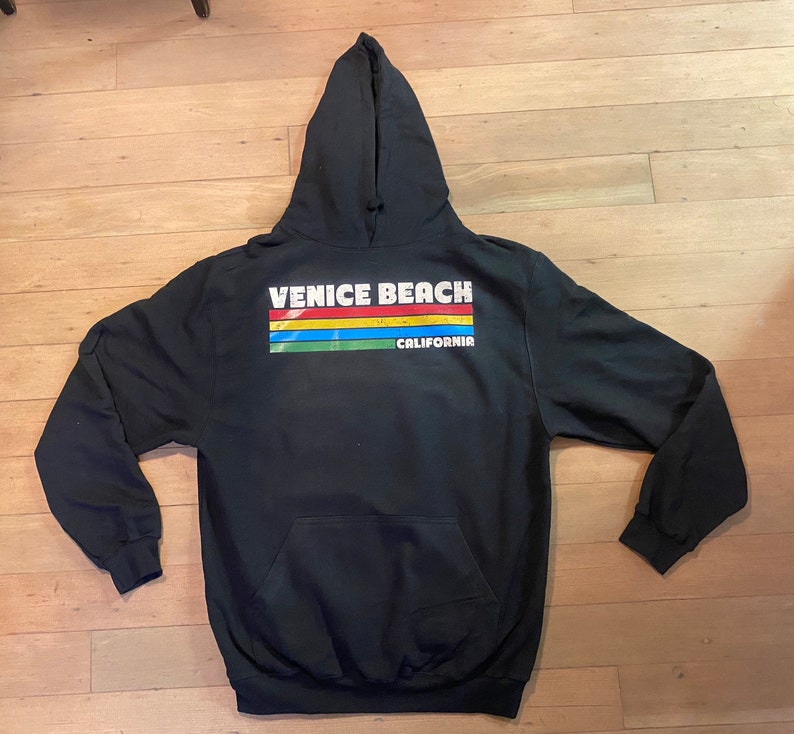 Puede incluir: Sudadera con capucha negra con un estampado gr&aacute;fico blanco que dice "Venice Beach California" con un dise&ntilde;o de rayas arco&iacute;ris.