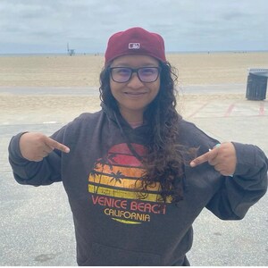 May include: A person wearing a dark gray hoodie with a vintage sunset graphic that says "Venice Beach California". The person is wearing a red baseball cap with a white logo and black framed glasses.
