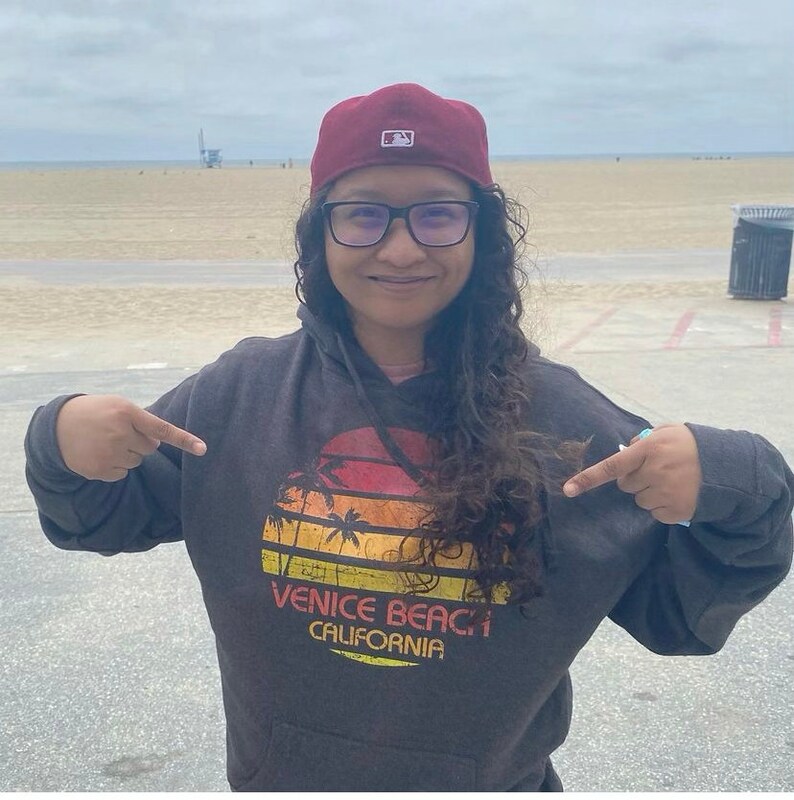 May include: A person wearing a dark gray hoodie with a vintage sunset graphic that says "Venice Beach California". The person is wearing a red baseball cap with a white logo and black framed glasses.
