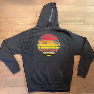 May include: A dark gray hooded sweatshirt with a vintage-style graphic of a sunset over palm trees and the text "Venice Beach California".