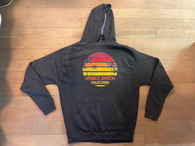 May include: A dark gray hooded sweatshirt with a vintage-style graphic of a sunset over palm trees and the text "Venice Beach California".