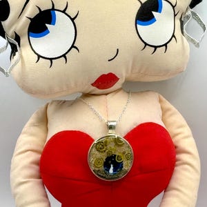 May include: A silver pendant necklace with a round, glass-covered charm featuring a whimsical design of a white rabbit under a starry night sky. The charm is set against a backdrop of gold and green floral elements, and is displayed on a red dress.