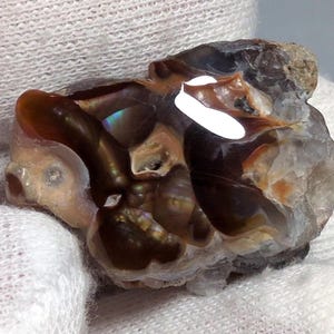 May include: A raw, uncut gemstone with a mix of brown, tan, and clear sections. The stone has a rough, natural texture and exhibits a play of colors, including iridescent flashes. The gemstone is held in a white glove, highlighting its natural beauty.