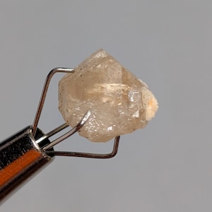 May include: A small, rough-cut, light-brown crystal is held by a silver-colored metal tweezer. The crystal has a slightly translucent appearance and a textured surface. The background is a solid, light gray.