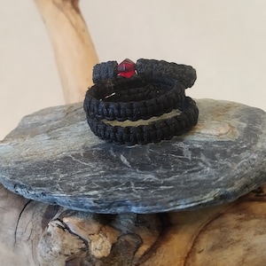 Puede incluir: Un brazalete de paracord negro con una cuenta de cristal roja. El brazalete está enrollado y sentado sobre una piedra gris.