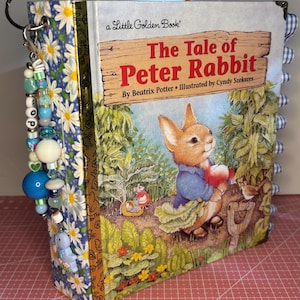 May include: A Little Golden Book titled "The Tale of Peter Rabbit" by Beatrix Potter, illustrated by Cyndy Szekeres. The book features a cover illustration of a rabbit holding an apple. The book's spine is decorated with blue and white daisy print fabric and a beaded charm.