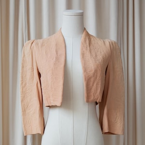 May include: A peach-colored bolero jacket with a textured, embossed pattern. The jacket has three-quarter sleeves and a shawl collar. It is displayed on a white dress form against a neutral background.