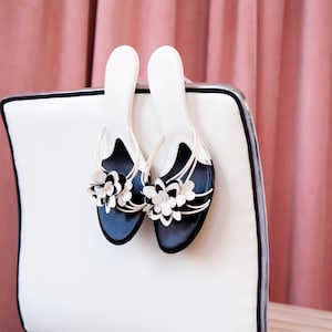 Op de afbeelding: Een paar zwarte sandalen met witte bloemenversieringen, rustend op een witte stoel met een zwarte rand.