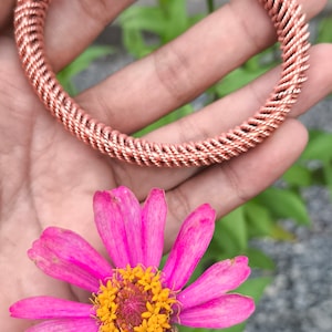 May include: A copper wire bracelet with a braided design. The bracelet is shown on a hand against a background of pink flowers and green foliage.